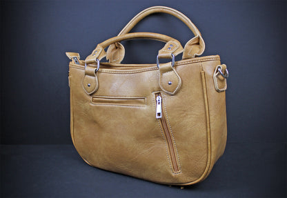 A tan faux-leather handbag with dual top handles, a front zippered pocket, and silver-tone hardware, photographed against a dark gray studio background.