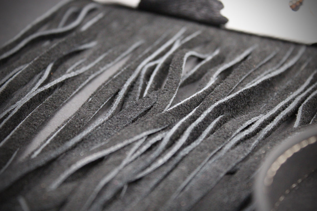 Close-up of a dark gray suede or leather fringe accessory with layered, wavy cut strips and a small metallic ring detail.