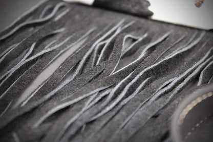 Close-up of a dark gray suede or leather fringe accessory with layered, wavy cut strips and a small metallic ring detail.