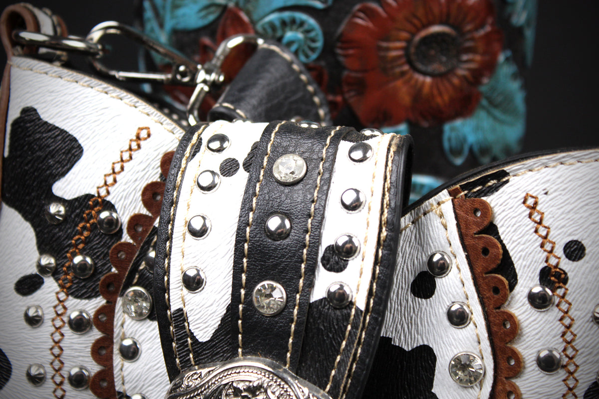 Close-up of a western-style handbag featuring a black and white cow-print leather body, silver-tone studs, chain straps, and a floral embossed background.