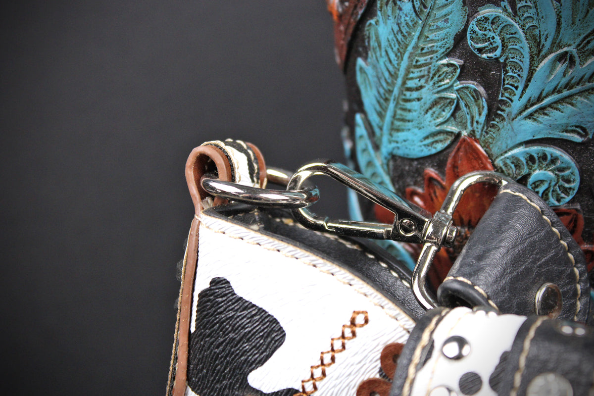 Close-up detail of a western-style leather bag or holster with a black-and-white cowhide patch, brown leather trim, silver metal clips, and turquoise-and-black tooled floral decoration on a dark background.