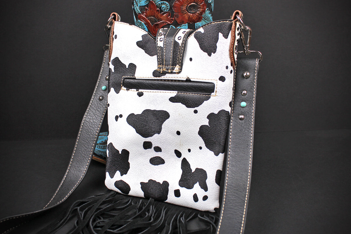 A western-style shoulder bag with a black and white cow print body, dark gray leather straps, and turquoise stud accents, displayed against a dark background.