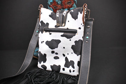 A western-style shoulder bag with a black and white cow print body, dark gray leather straps, and turquoise stud accents, displayed against a dark background.
