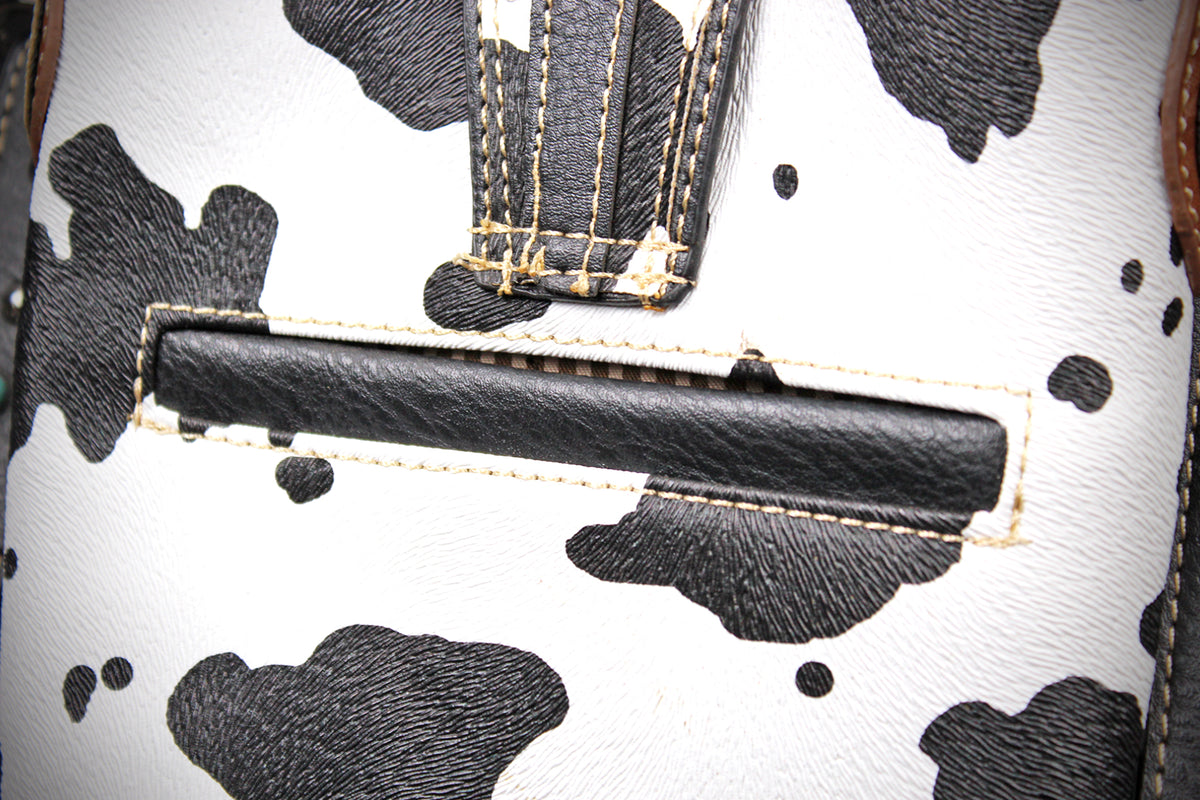 Close-up of a cow-print handbag showing a black leather horizontal slot detail on a white faux-leather background with black cow spots and contrast stitching.