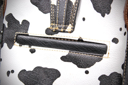 Close-up of a cow-print handbag showing a black leather horizontal slot detail on a white faux-leather background with black cow spots and contrast stitching.