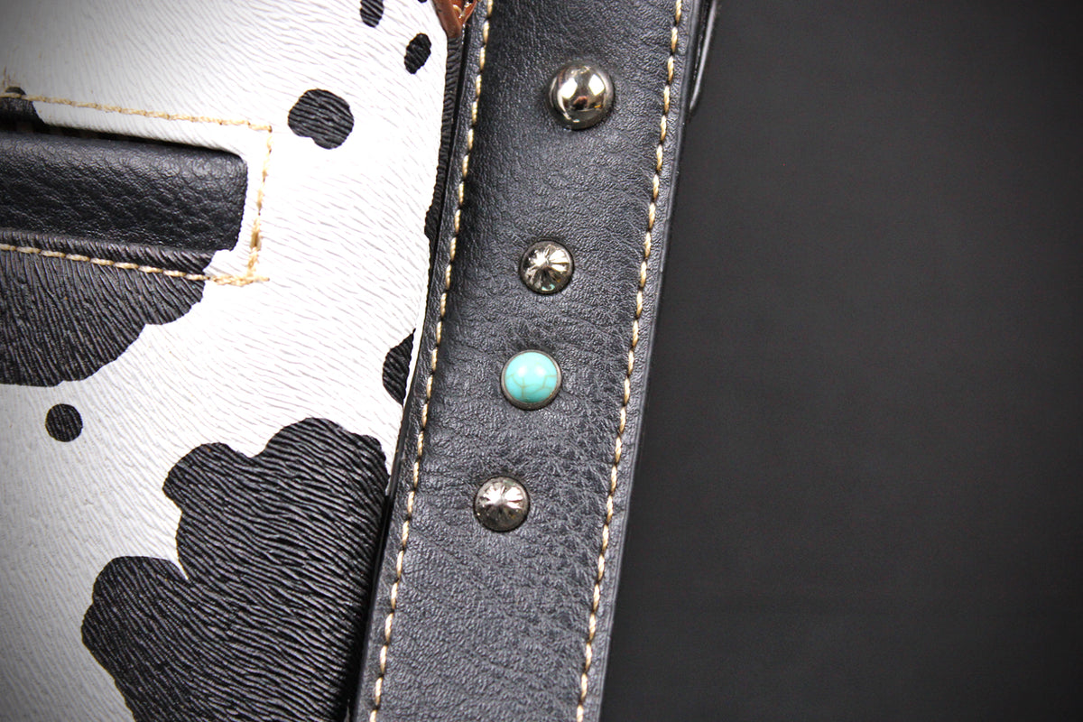 Close-up detail of a handbag showing a black and white cow-print leather panel, a black leather strap with silver studs, and a turquoise-colored accent stud on a dark gray background.