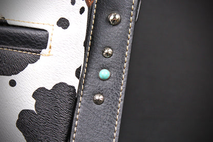 Close-up detail of a handbag showing a black and white cow-print leather panel, a black leather strap with silver studs, and a turquoise-colored accent stud on a dark gray background.