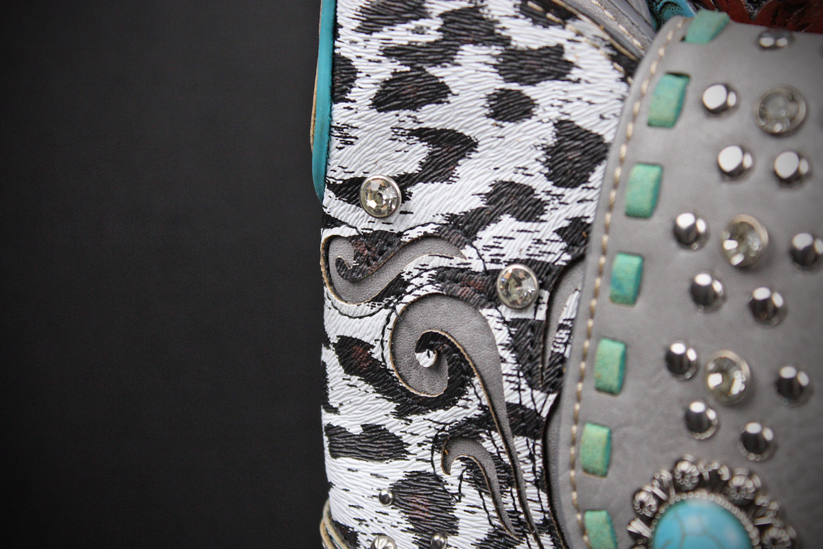 Close-up detail of a western-style boot showing a black and white cow-print side panel with silver conchos, a gray side panel with turquoise and silver studs, and a turquoise accent.