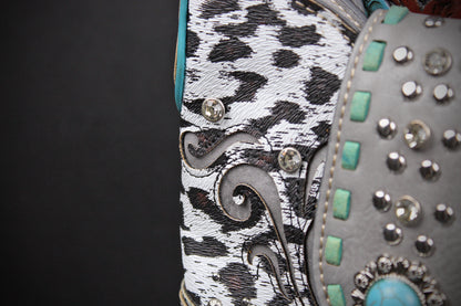 Close-up detail of a western-style boot showing a black and white cow-print side panel with silver conchos, a gray side panel with turquoise and silver studs, and a turquoise accent.