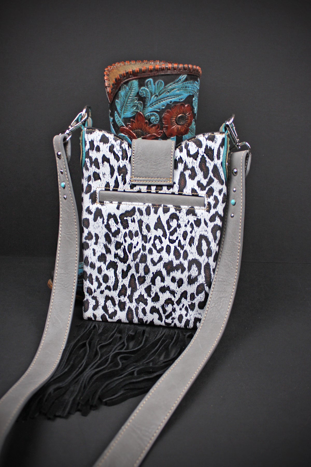 A western-style crossbody bag with a black and white leopard print body, black fringe, a gray strap, and a turquoise and brown tooled leather top flap, shown on a dark gray background.