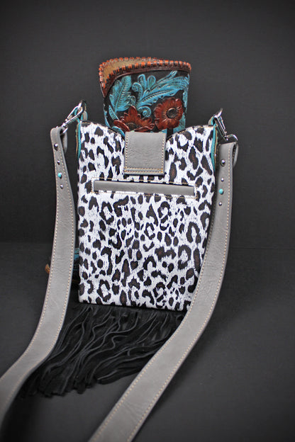 A western-style crossbody bag with a black and white leopard print body, black fringe, a gray strap, and a turquoise and brown tooled leather top flap, shown on a dark gray background.