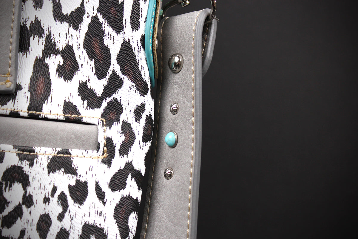 Close-up detail of a handbag showing a white and black leopard print fabric, a gray leather strap, silver-tone metal studs, and a turquoise accent bead against a dark background.
