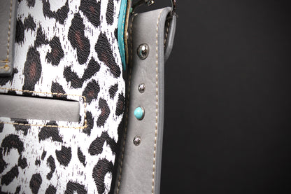 Close-up detail of a handbag showing a white and black leopard print fabric, a gray leather strap, silver-tone metal studs, and a turquoise accent bead against a dark background.