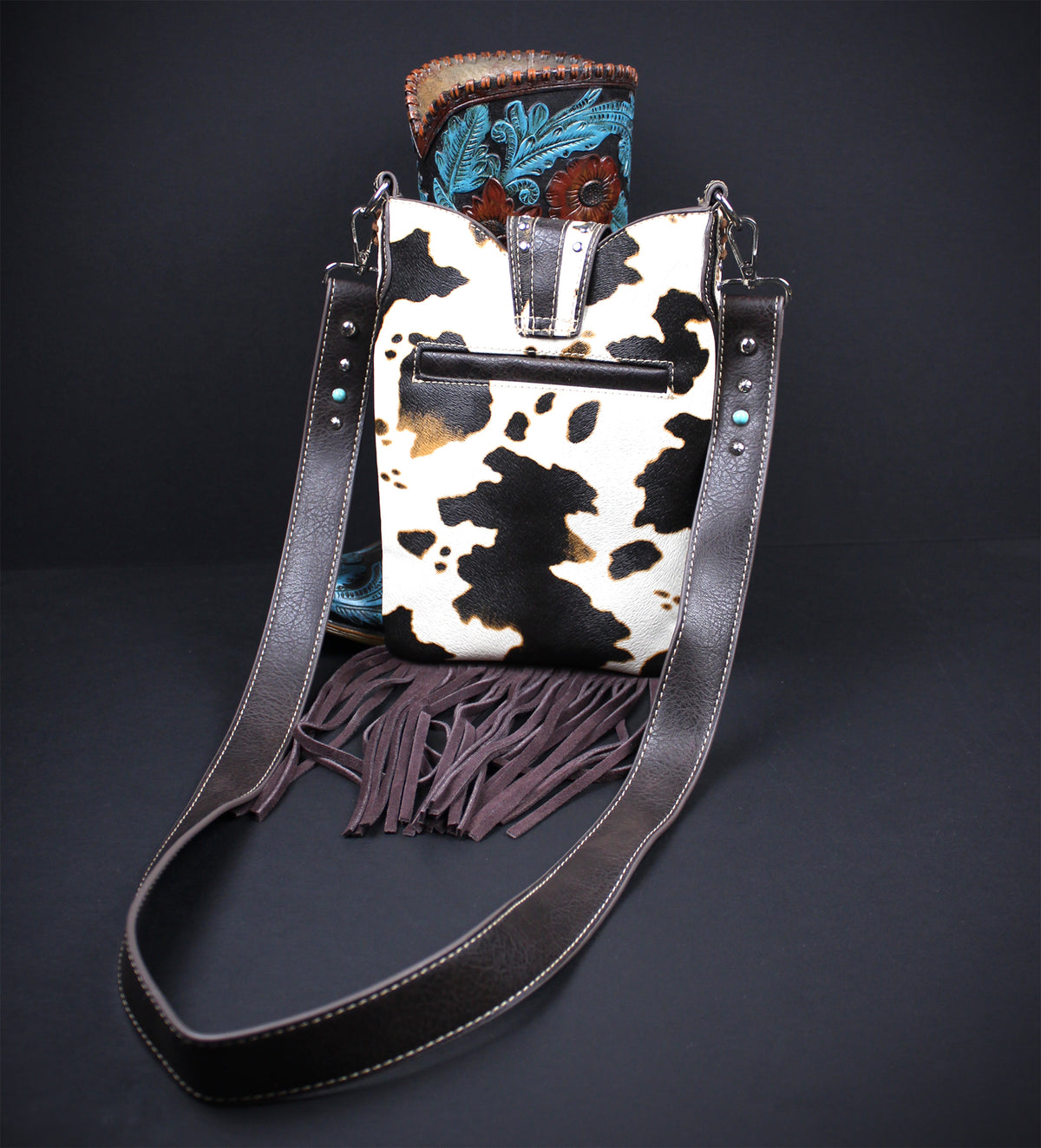 A western-style bucket bag with a black and white cowhide pattern body, dark brown leather fringe, and a long dark brown shoulder strap, displayed against a dark background with a boot tucked behind it.