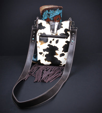 A western-style bucket bag with a black and white cowhide pattern body, dark brown leather fringe, and a long dark brown shoulder strap, displayed against a dark background with a boot tucked behind it.