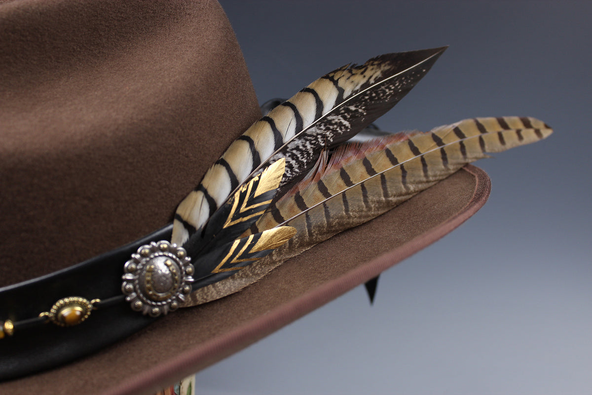 A close-up of a brown felt hat featuring a black band adorned with a large feathered ornament in black, beige, and gold tones, set against a soft gray gradient background.