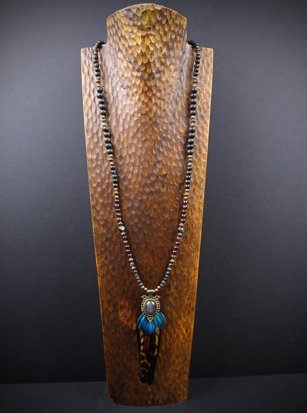 A long beaded necklace displayed on a textured wooden stand against a dark gray background. The necklace features dark brown beads, a bronze-toned metal connector, and a pendant with bright blue feathers and a brown-and-black tiger stripe pattern.