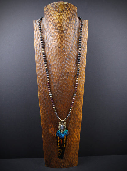 A long beaded necklace displayed on a textured wooden stand against a dark gray background. The necklace features dark brown beads, a bronze-toned metal connector, and a pendant with bright blue feathers and a brown-and-black tiger stripe pattern.