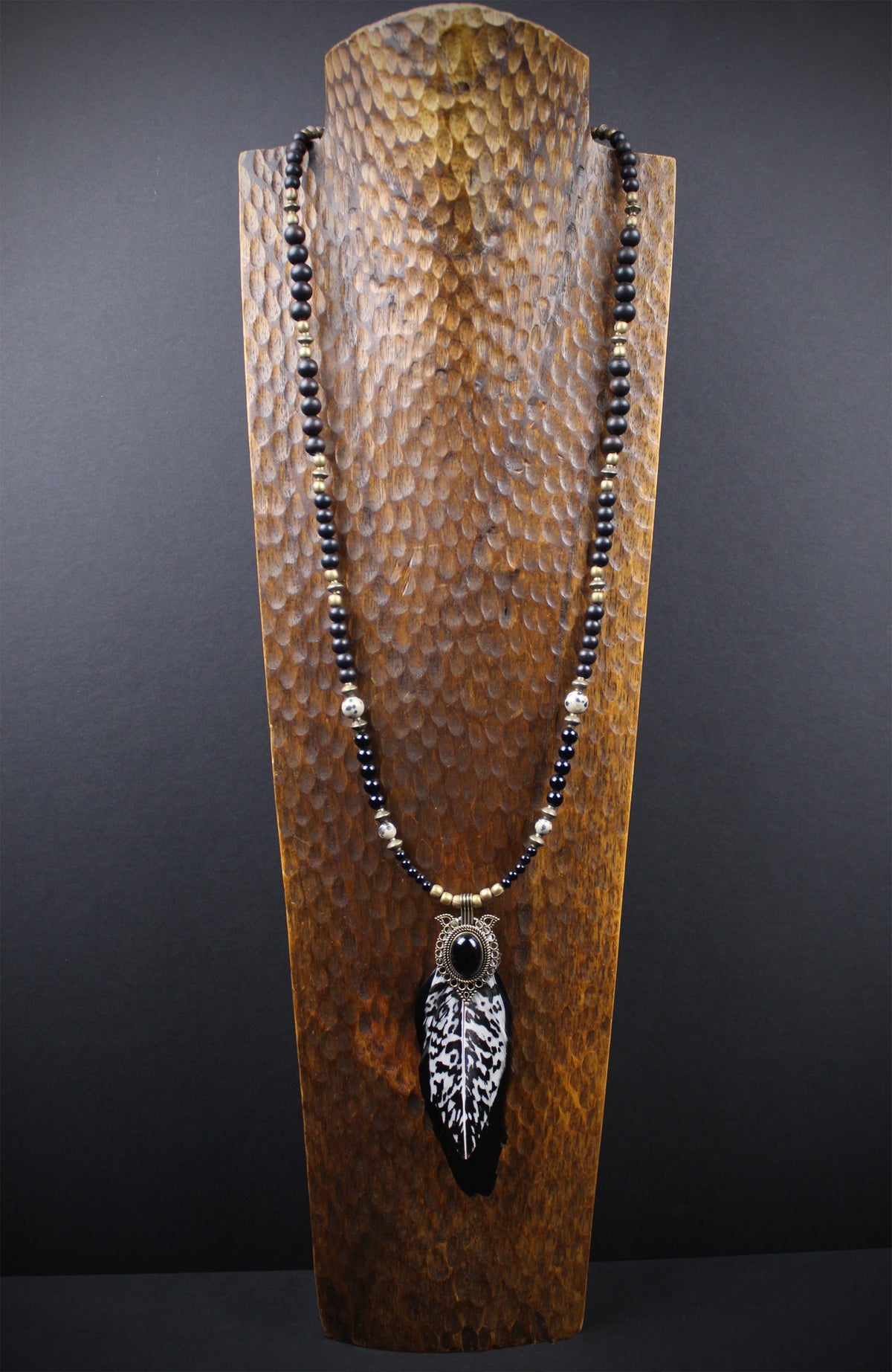 A beaded necklace with a large feather pendant displayed on a textured wooden stand against a dark gray background.
