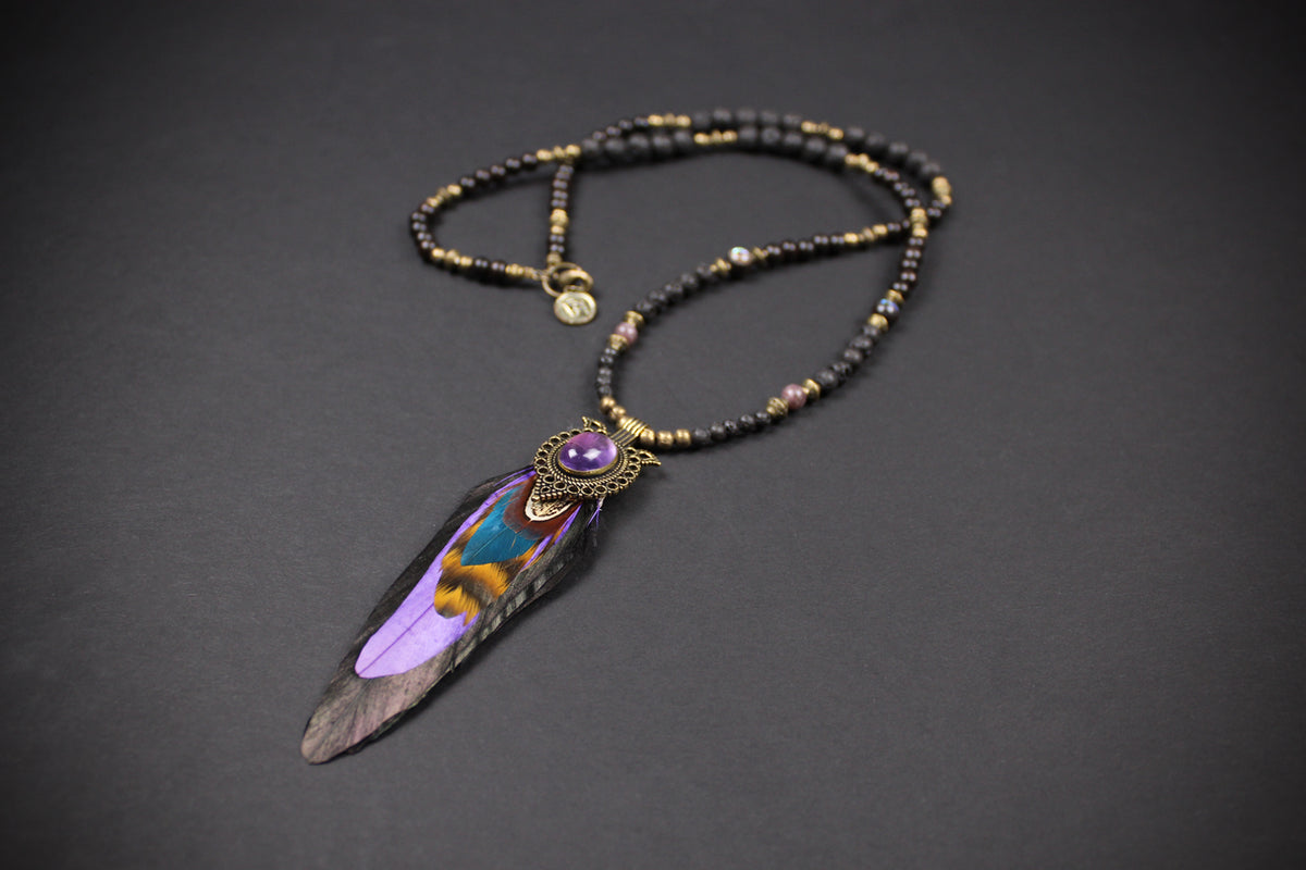 A beaded necklace with a large feather pendant, featuring black and gold beads, a purple stone accent, and a detailed purple, blue, and brown feather.