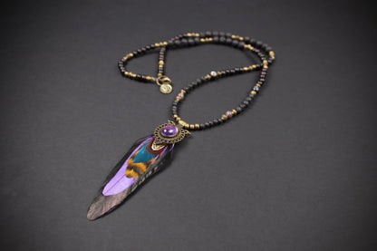 A beaded necklace with a large feather pendant, featuring black and gold beads, a purple stone accent, and a detailed purple, blue, and brown feather.