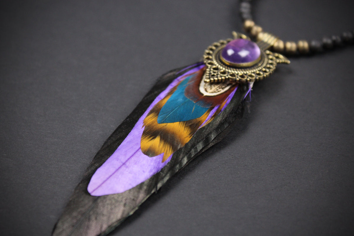 A close-up of a bohemian feather pendant necklace featuring a black feather with vibrant purple, blue, and gold iridescence, topped with an antique bronze filigree frame and a purple cabochon stone, on a dark gray background.