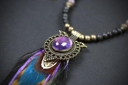 A close-up of a bohemian-style pendant necklace featuring a purple amethyst cabochon, ornate bronze filigree, a blue and purple feather, and a beaded chain on a dark background.