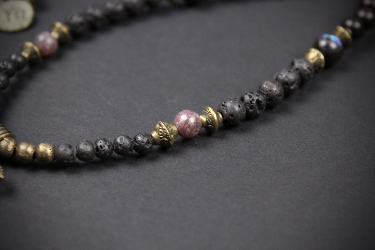 A beaded necklace featuring black lava stone beads, gold-toned spacer beads, and a central pink stone bead, displayed on a dark textured background.