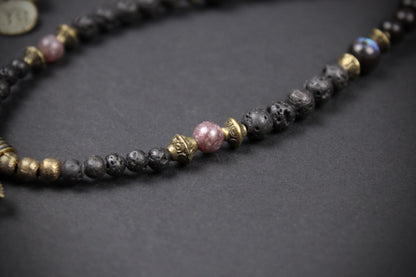 A beaded necklace featuring black lava stone beads, gold-toned spacer beads, and a central pink stone bead, displayed on a dark textured background.