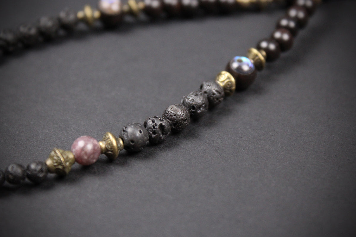 A close-up of a beaded necklace featuring dark brown lava stone beads, textured black beads, a pink stone bead, and antique gold metal spacers, set against a dark gray background.