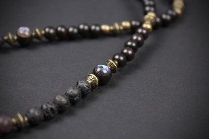 A close-up of a beaded necklace featuring dark brown wooden beads, textured black lava stone beads, and antique bronze metal spacers, with a central iridescent purple-blue bead accent, set against a dark gray background.