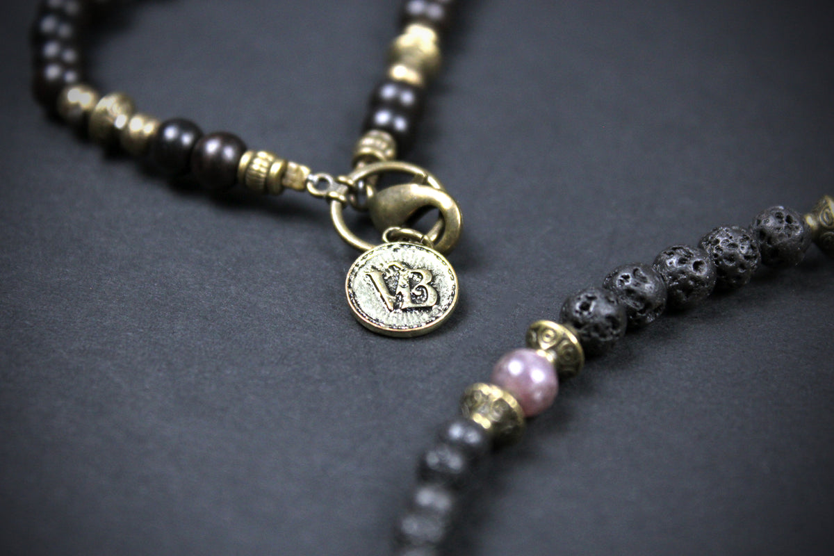 A close-up of a beaded necklace with dark brown and black beads, antique gold-toned spacers, a lobster clasp, and a round engraved coin charm, shown on a dark gray background.