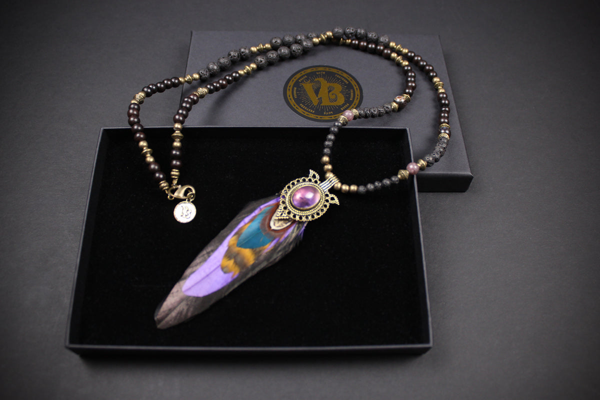 A beaded necklace with a large feather pendant displayed in an open black presentation box. The necklace features dark beads with metallic gold and silver accents, a small round charm, and a central ornate gold-toned connector holding a purple and iridescent feather.