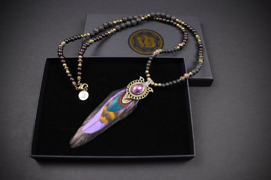 A beaded necklace with a large feather pendant displayed in an open black presentation box. The necklace features dark beads with metallic gold and silver accents, a small round charm, and a central ornate gold-toned connector holding a purple and iridescent feather.