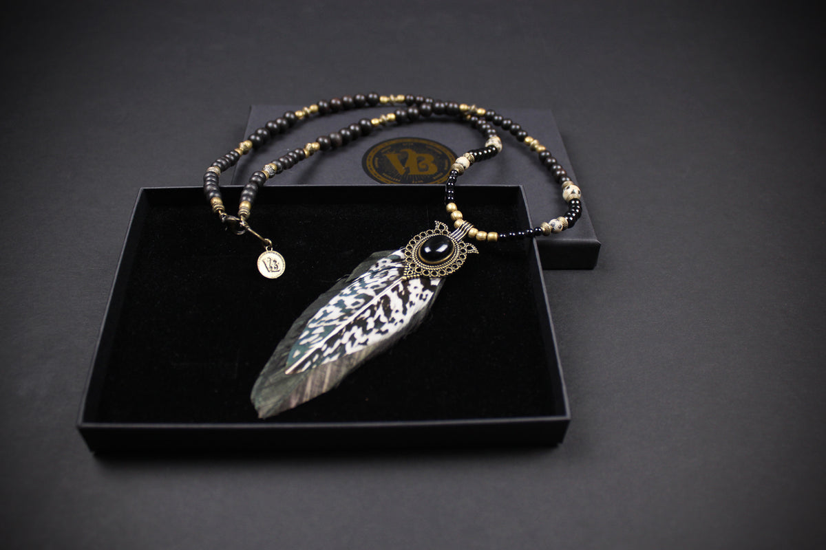 A silver-toned feather pendant necklace with a black and gold beaded chain, displayed in an open black presentation box on a dark background.