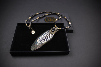 A silver-toned feather pendant necklace with a black and gold beaded chain, displayed in an open black presentation box on a dark background.