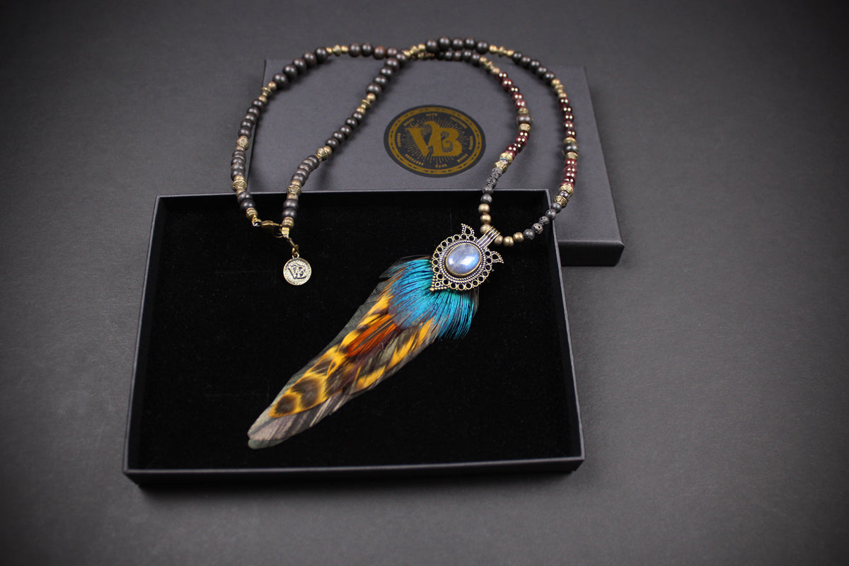 A beaded necklace with a large feather pendant displayed in an open black presentation box. The necklace features metallic bronze and brown beads, a silver-toned circular charm, and a central feather pendant with blue, orange, and brown feathering around a light blue stone.