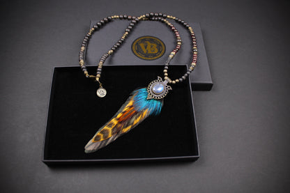 A beaded necklace with a large feather pendant displayed in an open black presentation box. The necklace features metallic bronze and brown beads, a silver-toned circular charm, and a central feather pendant with blue, orange, and brown feathering around a light blue stone.