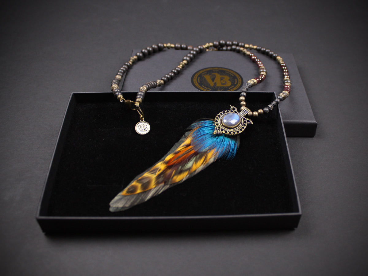 A beaded necklace with a large feather pendant displayed inside an open black presentation box. The necklace features dark metallic beads and a central feather with blue, orange, and black markings, set against a dark gray background.
