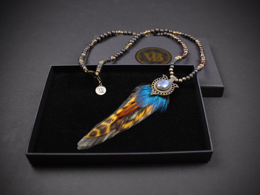 A beaded necklace with a large feather pendant displayed inside an open black presentation box. The necklace features dark metallic beads and a central feather with blue, orange, and black markings, set against a dark gray background.