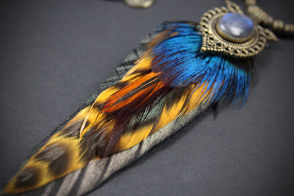 A close-up of a decorative feather pendant or ornament with a large oval blue stone in an ornate bronze-toned setting, attached to a long feather featuring blue, orange, yellow, black, and gray tones on a dark background.