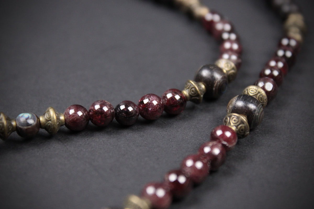 A close-up of a beaded necklace or mala-style strand with faceted dark red beads, metallic bronze spacer beads, and a few dark polished beads on a dark gray background.