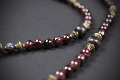 A close-up of a beaded necklace or mala-style strand with faceted dark red beads, metallic bronze spacer beads, and a few dark polished beads on a dark gray background.