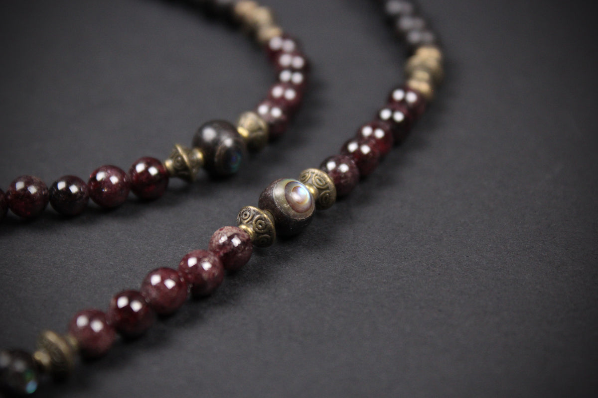A close-up of a beaded necklace with dark red polished gemstone beads, bronze-toned spacer beads, and one prominent iridescent white shell bead, set against a dark gray background.