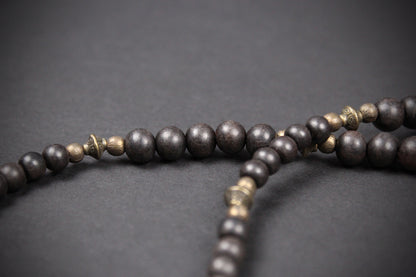 Dark brown wooden prayer beads with small metallic gold spacer beads, arranged on a dark gray background.