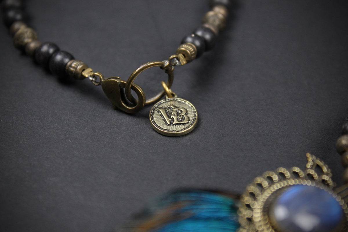 A close-up of a beaded necklace with dark round beads, antique gold spacer beads, a gold clasp, and a small round coin charm. A portion of a blue feather and ornate gold pendant are visible in the foreground on a dark textured background.