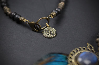 A close-up of a beaded necklace with dark round beads, antique gold spacer beads, a gold clasp, and a small round coin charm. A portion of a blue feather and ornate gold pendant are visible in the foreground on a dark textured background.