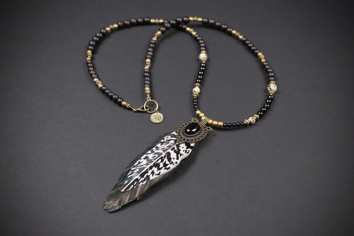 A beaded necklace with black and metallic gold beads, featuring a large feather pendant with black and white patterned detailing and a central black stone, displayed on a dark gray background.