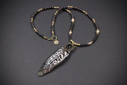 A beaded necklace with black and metallic gold beads, featuring a large feather pendant with black and white patterned detailing and a central black stone, displayed on a dark gray background.