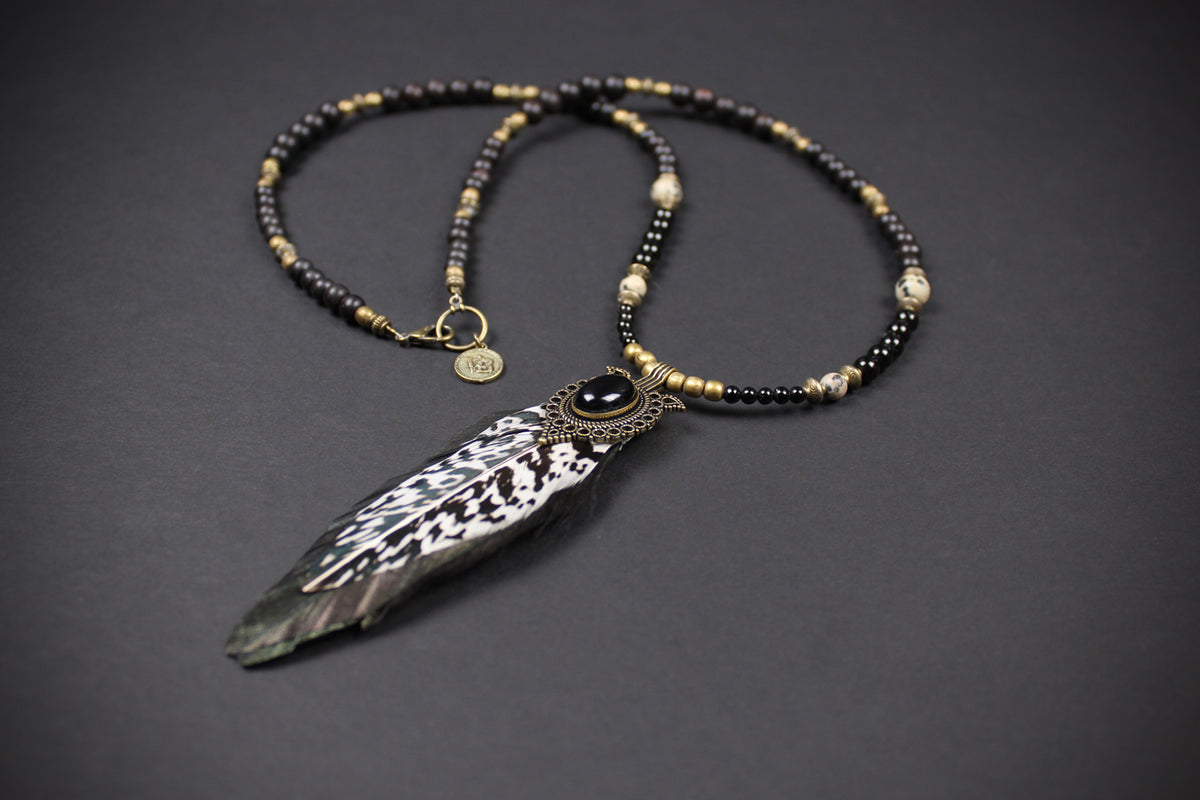 A bohemian-style beaded necklace featuring a large feather pendant, black and gold beads, and a central dark stone, displayed on a dark gray background.